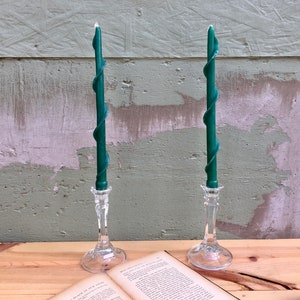 May include: Two green spiral candles in clear glass candlesticks. The candles are tall and slender, with a decorative spiral design. The candlesticks have a classic, elegant design. The background includes a wooden surface and a weathered green wall.