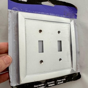 May include: A silver-colored decorative wallplate with two toggle switch openings. The wallplate is in a clear plastic package with the brand name "Amerelle" visible. The package also includes the words "Decorative Wallplate" and "Plaque murale décoratif".