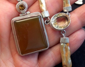 May include: A collection of handcrafted necklaces featuring various gemstones and natural elements. The necklaces include a rectangular brown stone pendant, a citrine oval pendant, and a strand of amber-colored beads. The jewelry is set in silver.