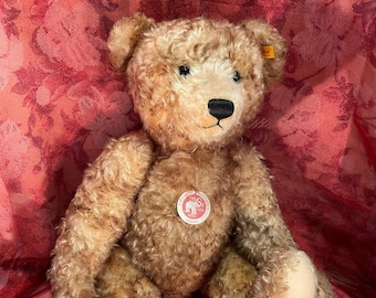 May include: A light brown curly mohair teddy bear with a black stitched nose and mouth. The bear features a round white chest tag with a red elephant logo and text, a small yellow ear tag, and light beige felt paw pads on its feet.