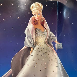 Puede incluir: Muñeca rubia con un vestido plateado con adornos brillantes y un chal a juego. La muñeca sostiene un pequeño bolso cuadrado. El fondo es azul oscuro con formas blancas en forma de estrella.