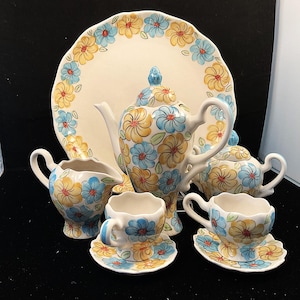 May include: A ceramic tea set with a floral pattern in yellow and blue. The set includes a teapot, sugar bowl, creamer, two teacups, saucers, and a serving platter. The tea set is on a black surface.