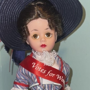 May include: A doll dressed as a suffragette, wearing a blue and white striped dress with a red sash that reads "Votes for Women." The doll has a large blue hat and red shoes. The doll is part of the "Through the Decades Collection."