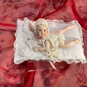 May include: A porcelain baby doll wearing a lace-trimmed bonnet and romper, lying on a white pillow with lace edging. The doll has a small teddy bear and a rattle. The background is a red floral fabric.