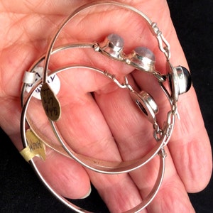 May include: Three silver hoop bracelets with gemstone accents. The top bracelet has two round, light blue gemstones. The middle bracelet has a round, black gemstone. The bottom bracelet is plain.