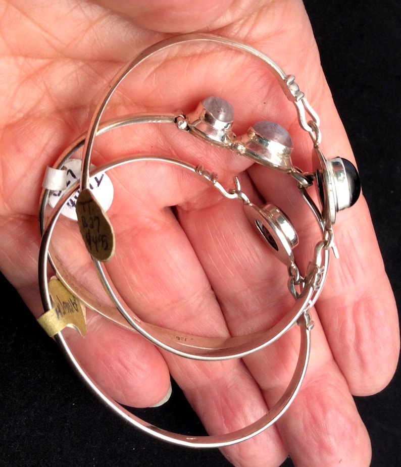May include: Three silver hoop bracelets with gemstone accents. The top bracelet has two round, light blue gemstones. The middle bracelet has a round, black gemstone. The bottom bracelet is plain.