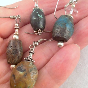 Pode incluir: Três pares de brincos com ganchos de prata e pedras de forma oval. As pedras são uma mistura de marrom, verde e azul.