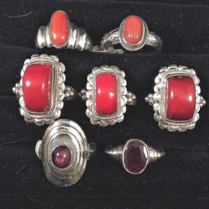 May include: A collection of silver rings with various red and maroon gemstones. The rings feature oval, rectangular, and round stones, set in ornate silver bands. The gemstones are a vibrant red and maroon color.