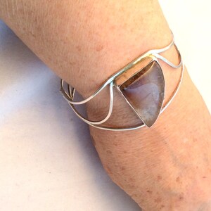May include: A silver bracelet with a triangular shaped gemstone set in the center. The gemstone is a light brown color with a hint of purple.