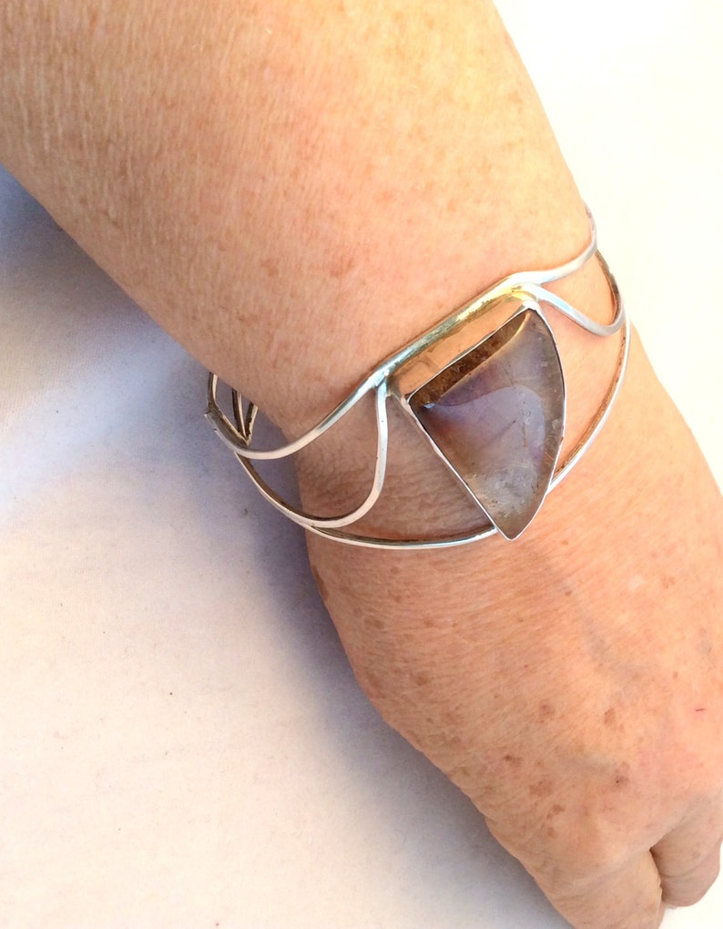 May include: A silver bracelet with a triangular shaped gemstone set in the center. The gemstone is a light brown color with a hint of purple.