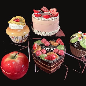 May include: A collection of decorative trinket boxes shaped like desserts and a red apple. The desserts include a cupcake, a cake with strawberries, a heart-shaped cake, and a cupcake with kiwi slices. The apple is red with a green stem.