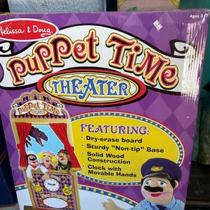 May include: A purple Melissa & Doug "Puppet Time Theater" box. The box features a puppet stage with a clock, puppets, and the text "Puppet Time Theater". Additional text includes "Dry-erase board", "Solid Wood Construction", and "Clock with Movable Hands".