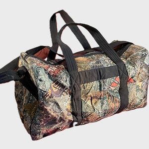 May include: A quilted duffel bag featuring a multi-colored paisley pattern in shades of green, brown, and orange. The bag includes black fabric handles and a matching black shoulder strap, with a rectangular base and a zippered top closure.