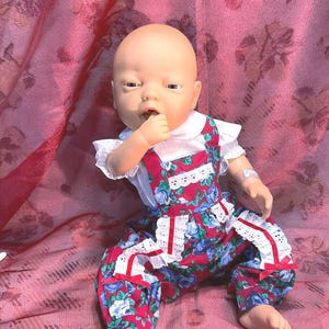 May include: A doll with a bald head and a white collared shirt with ruffled sleeves. The doll wears red overalls with a blue floral pattern and white lace trim. The doll is sitting on a pink floral patterned fabric.