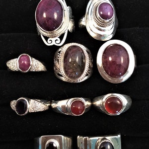 Pode incluir: Uma coleção de anéis de prata com várias configurações de pedras preciosas. Os anéis apresentam pedras ovais, redondas e retangulares em tons de vermelho e roxo. Alguns anéis têm detalhes intrincados em prata, enquanto outros têm um design de banda simples.