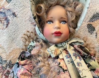 May include: A porcelain doll with blonde, curly hair and blue eyes. The doll wears a floral print dress and a matching bonnet tied with a bow. The dress features pink, purple, and green flowers.