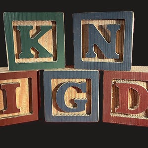 May include: Five wooden alphabet blocks spelling the word KIND. The blocks are painted in various colors: green, blue, and red. Each block has a letter cut out, revealing the natural wood grain.