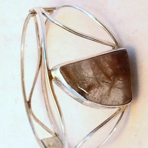 May include: A silver bangle bracelet with a large, clear, faceted gemstone set in the center. The gemstone has a brown and white color.