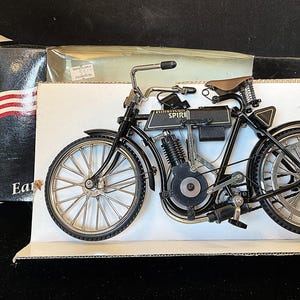 May include: A black and silver vintage motorcycle model with a brown seat. The model is displayed on a white surface with a box in the background that has the American flag and the words "U.S.A. 1903 American Spirit."