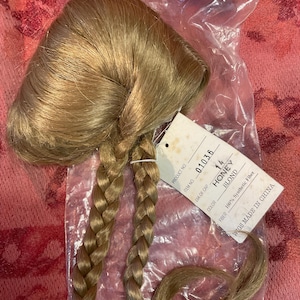 May include: A honey blonde doll wig with two braided pigtails, packaged in a clear plastic bag. The wig is labeled "Honey Blond" and made of 100% synthetic fiber. The packaging includes the brand name "Dollspari".