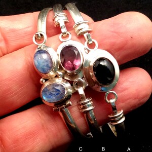 May include: Three silver bangle bracelets with gemstone charms. The charms are a light blue moonstone, a dark purple amethyst, and a black onyx. The bracelets are labeled A, B, and C.