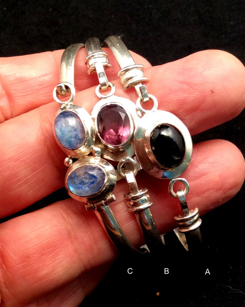 May include: Three silver bangle bracelets with gemstone charms. The charms are a light blue moonstone, a dark purple amethyst, and a black onyx. The bracelets are labeled A, B, and C.