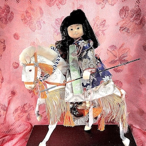 May include: A doll with black hair dressed in a blue and white patterned garment, mounted on a white horse with orange tassels and a black harness. The figure holds a long, thin black staff and is positioned on a rectangular black base.