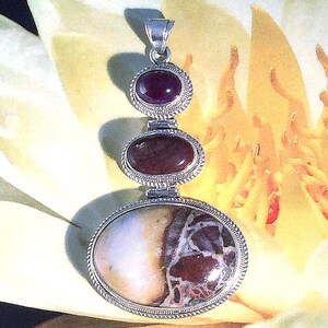 May include: A silver pendant with three oval-shaped stones. The top stone is a deep red, the middle is a darker brown, and the bottom is a cream and brown patterned stone. The pendant is set against a yellow and white background.