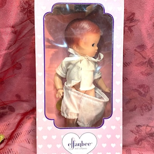 May include: A vintage Effanbee doll in its original box. The doll has red hair, a white shirt, and white shorts. The box is white with a pink heart pattern and the Effanbee logo.