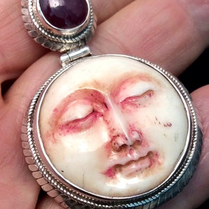 Pode incluir: Um pingente de prata com um rosto esculpido e uma gema roxa. O rosto é esculpido em detalhe e tem os olhos fechados. O pingente é rodeado por uma borda de prata.