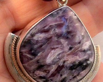 May include: A silver pendant with a large, purple, irregularly shaped gemstone. The pendant is set in a silver frame with a rope-like border. The gemstone has a swirling, marbled pattern.