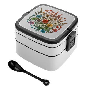 May include: A white, two-tiered lunch box with a black trim and a black spoon. The lid features a colorful floral design with various flowers. The lunch box has a black clasp on the side to secure the lid.