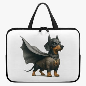 Op de afbeelding: Een witte laptoptas met zwarte rand en handvatten. De tas heeft een cartoonillustratie van een teckel verkleed als superheld, compleet met cape, masker en embleem. De hond is zwart en bruin.