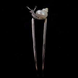 May include: A silver hair fork with a snail design. The snail has a pearlescent shell and detailed antennae. The fork's tines are long and slender, creating a decorative hair accessory.