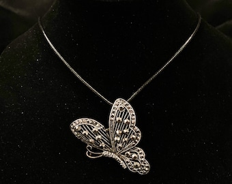 Puede incluir: Un collar con un colgante de mariposa de plata con diamantes de imitación negros en un cordón negro.