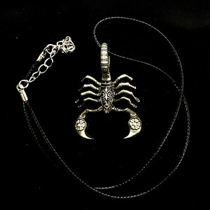 May include: A silver scorpion pendant necklace with a black cord. The scorpion has intricate details and a floral design on its tail.