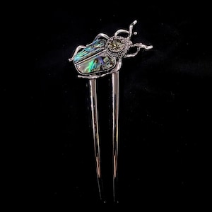 May include: A silver hair pin with a beetle design. The beetle's body is inlaid with iridescent abalone shell, displaying green, blue, and purple hues. The pin has two long, parallel prongs.