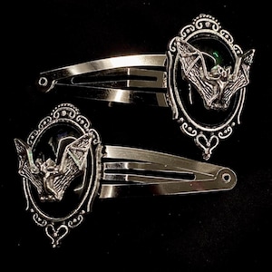 May include: Two silver-toned metal hair clips featuring an oval frame with an ornate filigree border. Each clip contains a black oval inlay centered with a three-dimensional silver bat figure with outstretched wings, attached to a snap-style barrette.