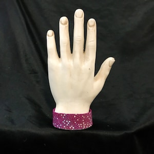 May include: A white ceramic hand with a pink and white patterned bracelet. The hand is raised with fingers spread apart.