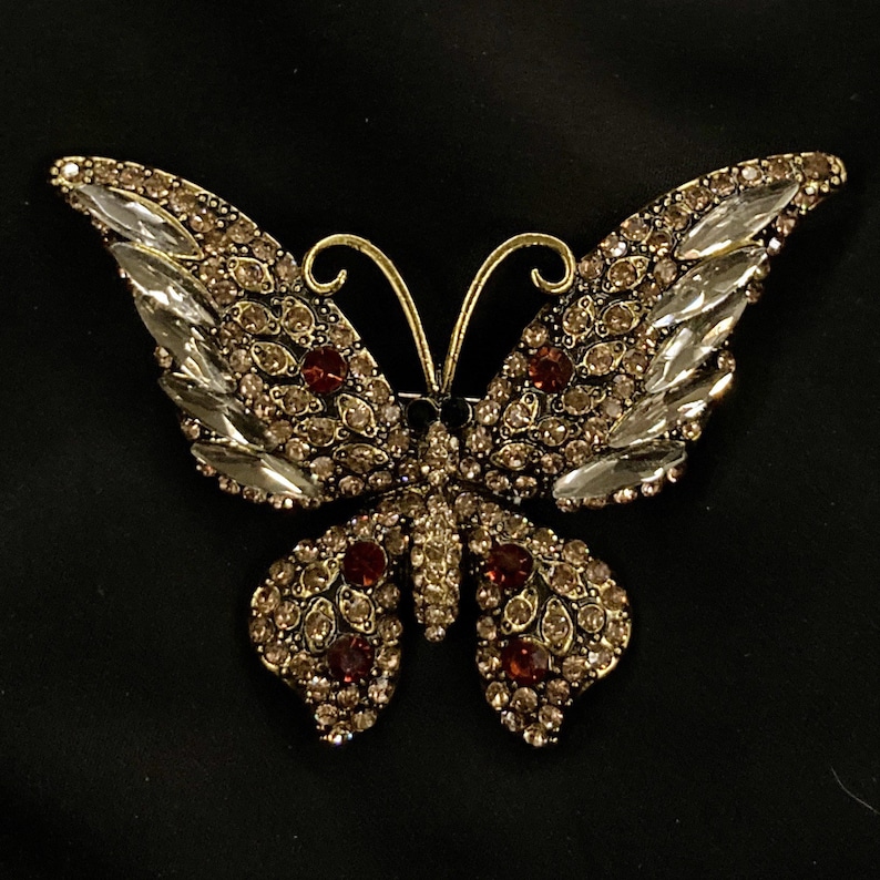 Puede incluir: Un broche de mariposa de oro con diamantes de imitaci&oacute;n transparentes y marrones. La mariposa tiene un dise&ntilde;o detallado con alas intrincadas y un cuerpo.