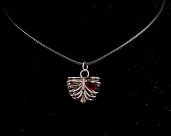 Puede incluir: Un collar con un colgante de caja torácica plateada con una gema roja en el centro, colgando de un cordón negro.