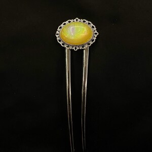 May include: A silver hair pin with an oval, yellow-colored stone. The stone is surrounded by a decorative, filigree metal frame. The pin has two long, parallel prongs.