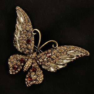 Puede incluir: Un broche de mariposa de tono dorado con diamantes de imitaci&oacute;n transparentes y marrones. Las alas de la mariposa est&aacute;n extendidas y el cuerpo est&aacute; decorado con dise&ntilde;os intrincados.