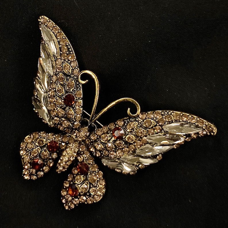 Puede incluir: Un broche de mariposa de tono dorado con diamantes de imitaci&oacute;n transparentes y marrones. Las alas de la mariposa est&aacute;n extendidas y el cuerpo est&aacute; decorado con dise&ntilde;os intrincados.