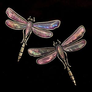 May include: Two dragonfly brooches with iridescent purple and silver wings and bodies. The brooches have a silver-tone metal frame and are set against a black background. The dragonfly jewelry is a beautiful accessory.
