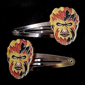 Puede incluir: Dos horquillas plateadas, cada una adornada con un diseño de rostro colorido. Los rostros presentan detalles amarillos, rojos y negros, con una expresión severa. Las horquillas están sobre un fondo negro.