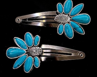 May include: Two silver-toned hair clips, each adorned with a turquoise flower design. The flower petals are oval-shaped, and the center has a starburst pattern. The clips are set against a black background.