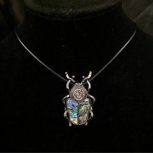 Puede incluir: Un collar con colgante de escarabajo plateado. El colgante presenta un diseño detallado de escarabajo con detalles iridiscentes de concha de abulón y un centro de cristal brillante. El collar cuelga de un cordón negro.