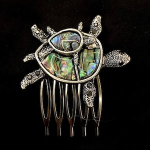Puede incluir: Peine de pelo plateado con un diseño de tortuga de nácar. La tortuga tiene detalles negros y una concha iridiscente.