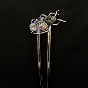 May include: Silver hair pin with a beetle design. The beetle's body is inlaid with iridescent abalone shell, and a green gemstone accents the head. The pin has two long, parallel prongs.